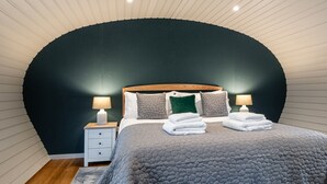 Un dormitorio con techo curvo, una cama con colcha gris, dos mesitas de noche con lámparas y un cabecero de madera.