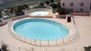 A large outdoor swimming pool with a surrounding fence, a smaller adjacent pool, and a building in the background. - Two-room flat, Le Grau du Roi (Le Grau-du-Roi)