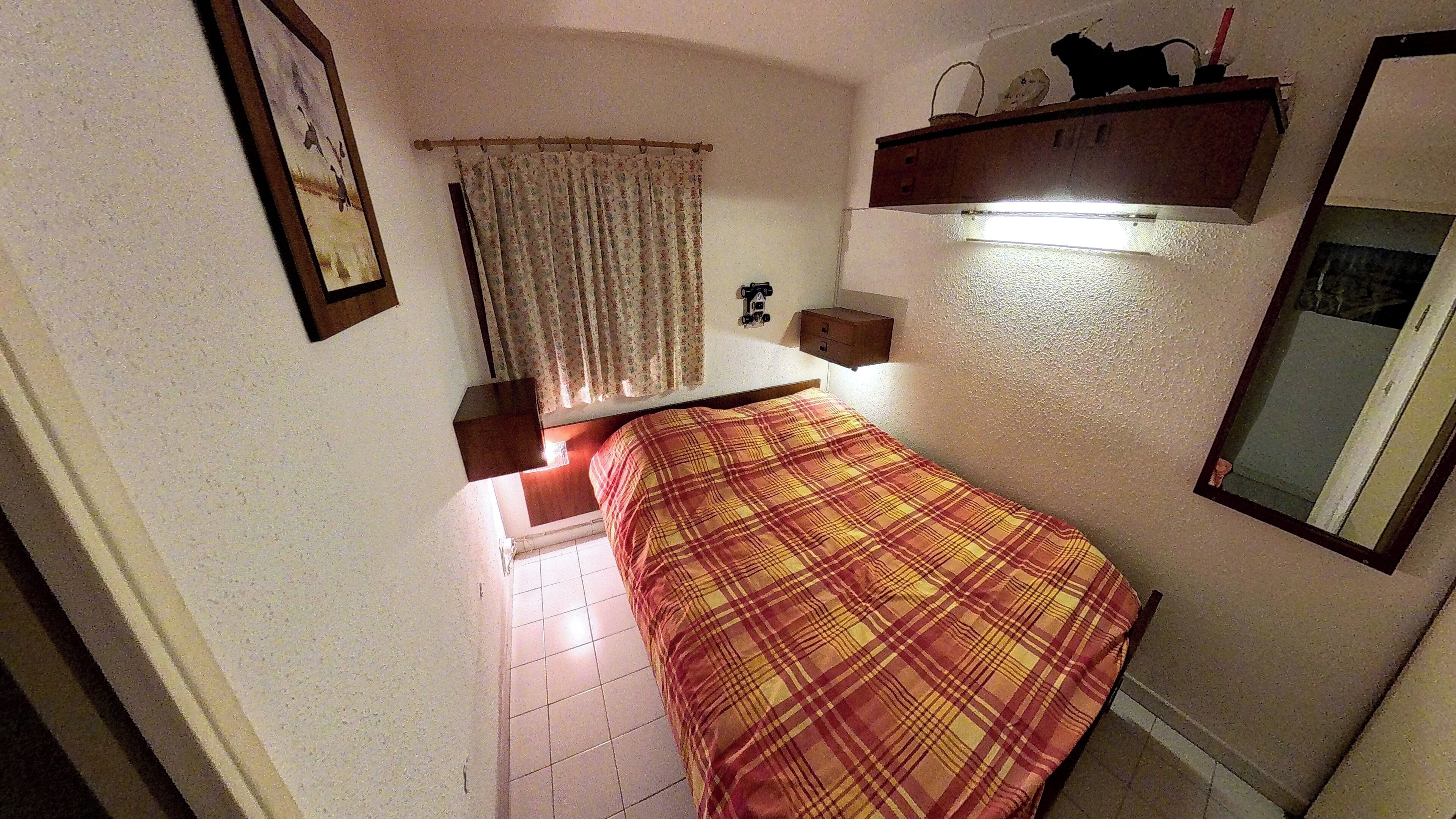 Um quarto pequeno e estreito, com uma cama, uma janela com cortinas, uma prateleira com objetos e uma luminária fixada na parede.