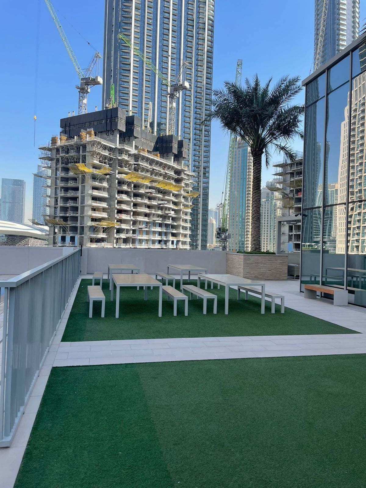 A rooftop terrace with artificial grass and white tables and benches, surrounded by high-rise buildings and construction cranes.