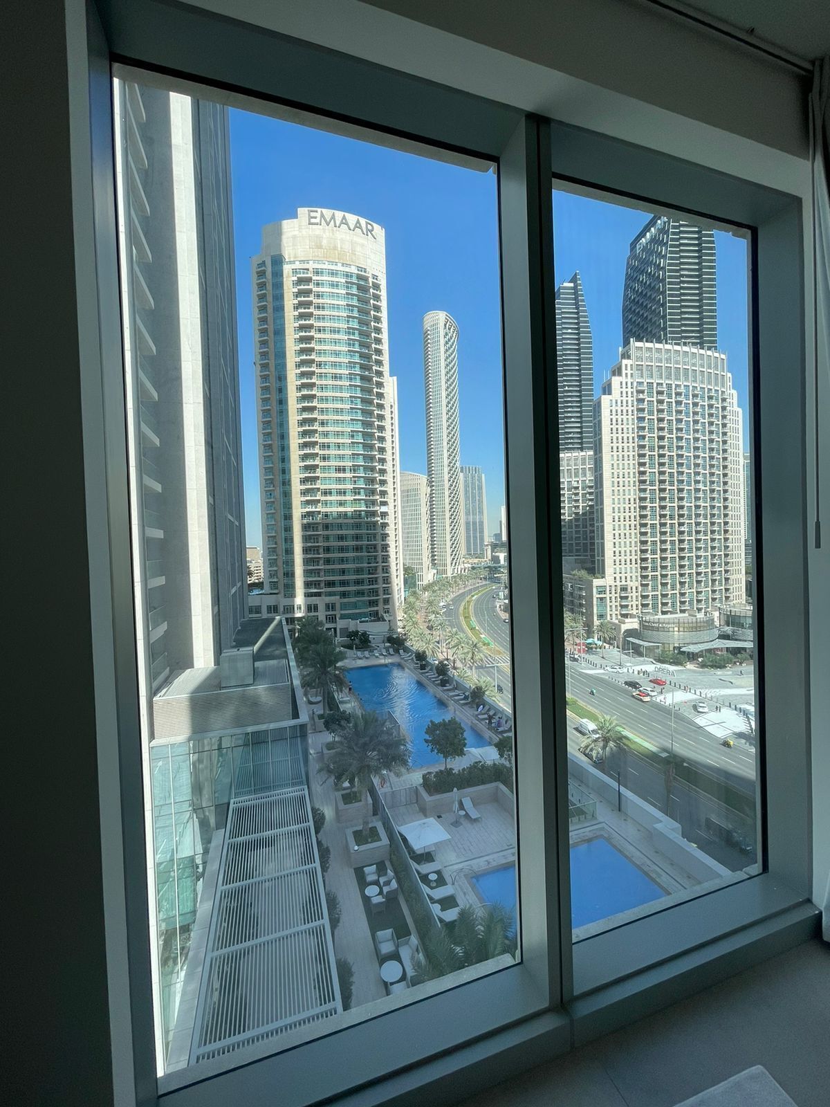 A view from a high-rise window overlooking a cityscape with modern buildings, including a prominent EMAAR building, and a landscaped area with a pool.