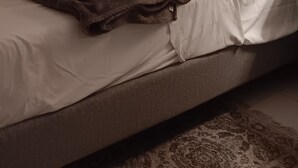 A bedroom with a bed, a brown headboard, a white pillow, a red decorative pillow, a brown blanket, and a patterned rug.