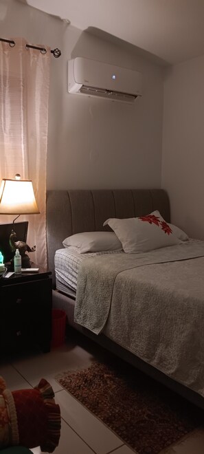 A bedroom with a bed, a nightstand, a lamp, and an air conditioning unit. - Los Angeles house. (Carolina)