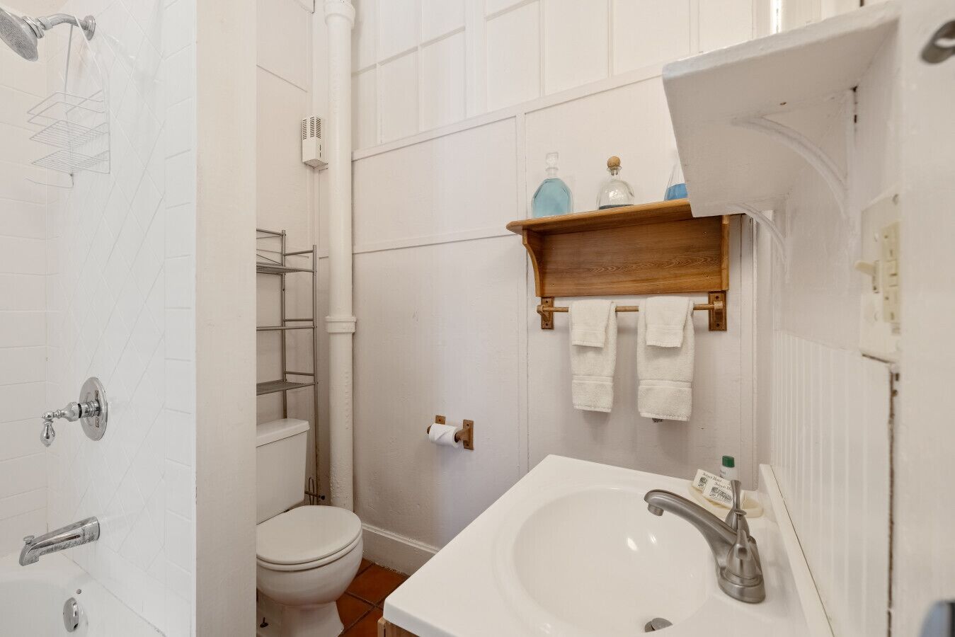 Un baño con lavamanos, inodoro y ducha blancos. Hay un estante de madera sobre el lavamanos con una toalla colgada.
