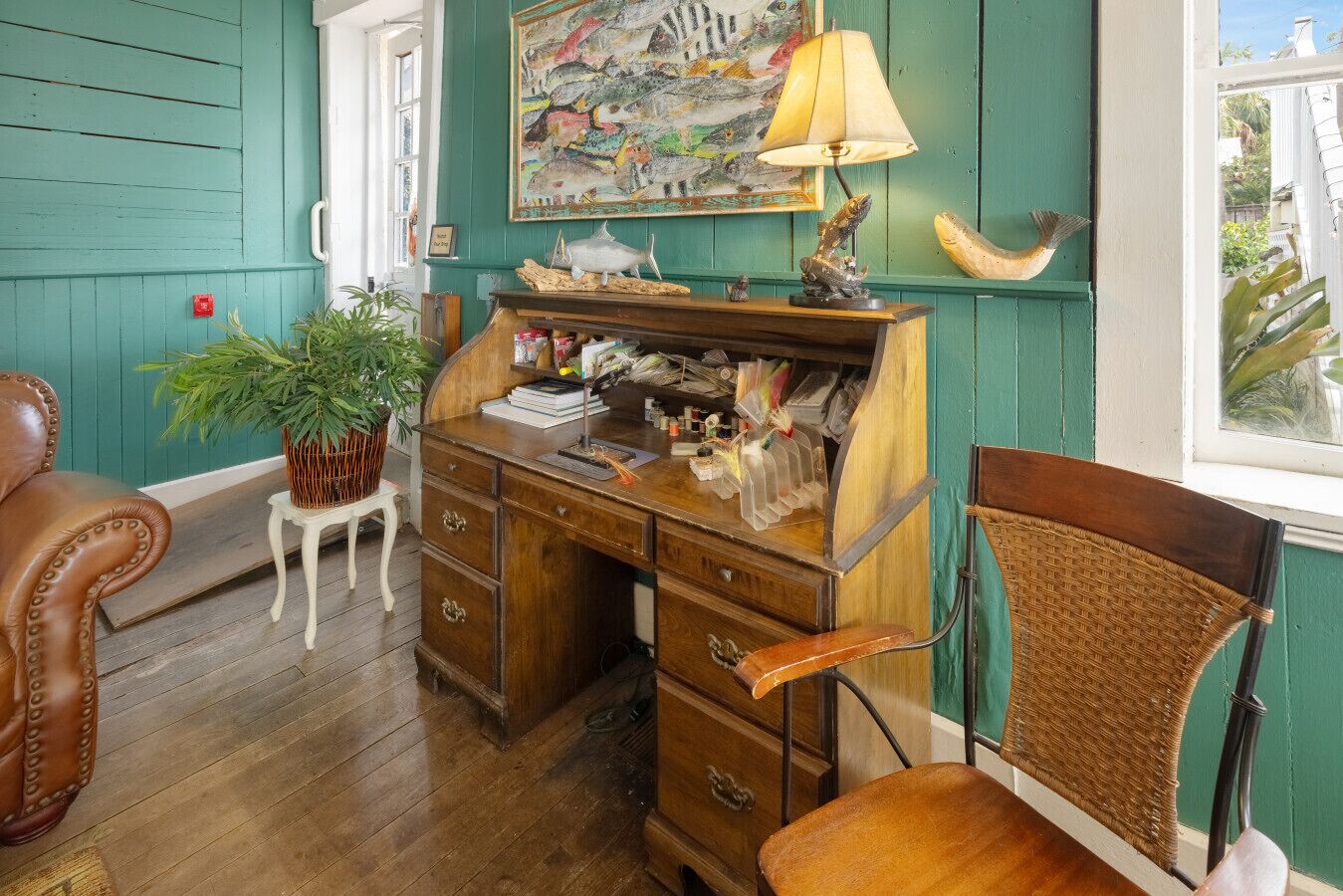 A room with teal walls, a wooden desk with drawers, a wicker chair, and a potted plant.