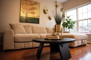 Un salon avec un canapé beige, une table basse ronde noire, une grande fenêtre et un tableau représentant un arbre.