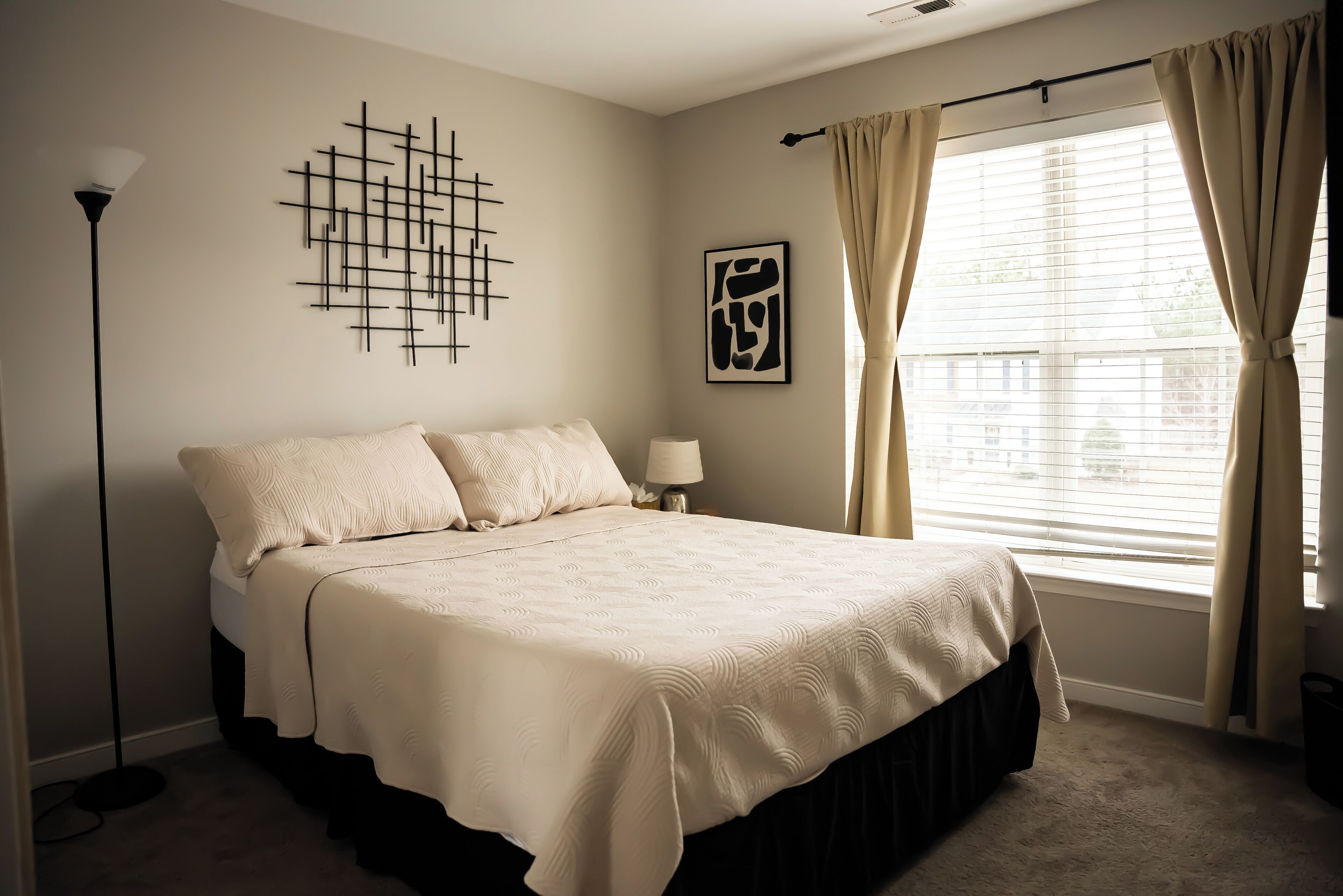 A bedroom with a large bed, a floor lamp, a wall art piece, and a window with curtains.