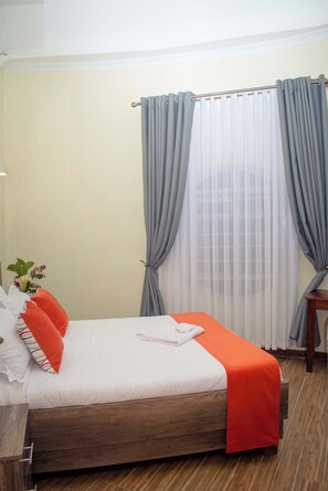 A neatly made bed with a white and orange bedspread, grey curtains, and a wooden floor. - Turaco Intercounty Resort Limited  (Kisumu)