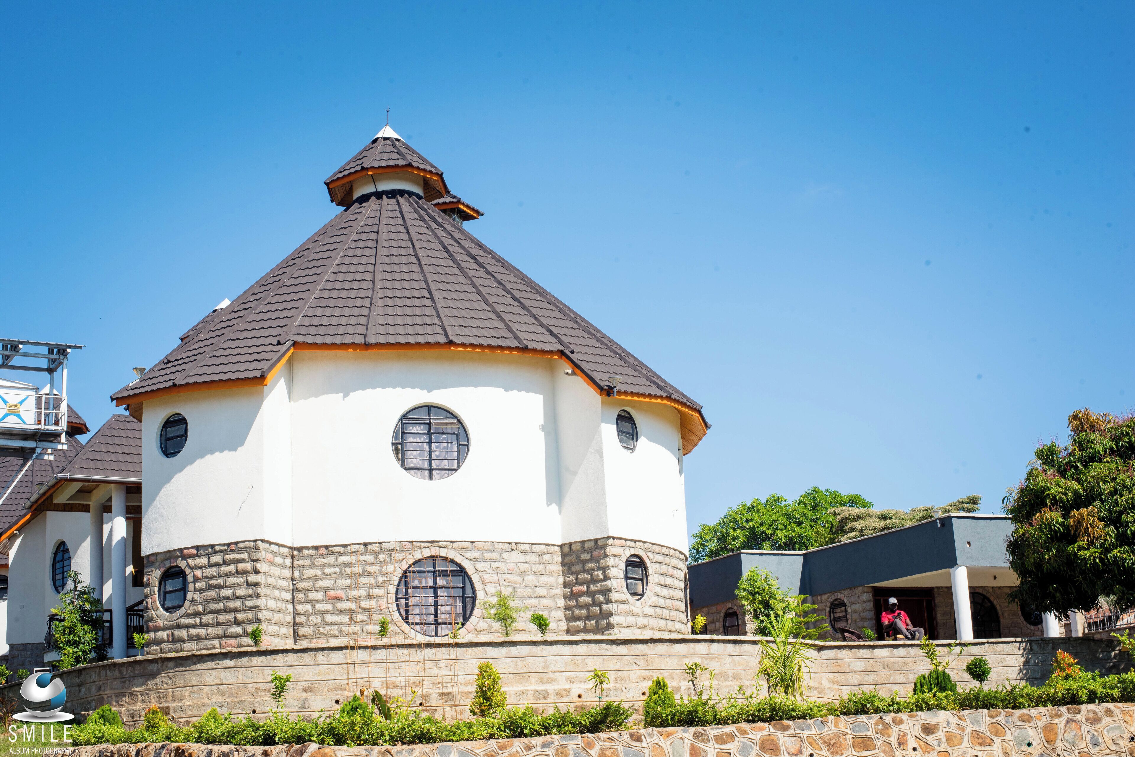 A white building with a conical roof and multiple windows, surrounded by stone walls and greenery.