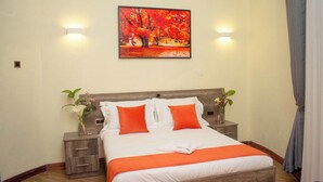 A neatly made bed with white and orange bedding, two bedside tables with lamps, and a painting of autumn trees on the wall. - Turaco Intercounty Resort Limited  (Kisumu)