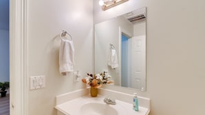 Un baño con lavamanos blanco, un espejo, un perchero para toallas y un florero con flores.