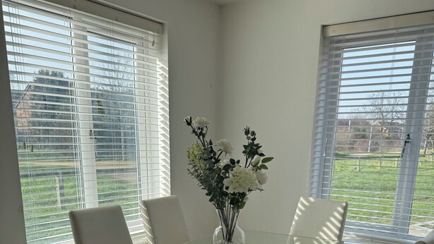 Un comedor moderno con una mesa de vidrio rodeada de sillas de cuero blanco, un jarrón con flores y ventanas amplias con persianas.
