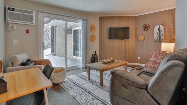 A living room with a sofa, a coffee table, and a TV mounted on the wall. There is a sliding glass door leading to a balcony. - Jelinek Hideaway Unit 5 - Modern 2BR Apartment (Schofield)