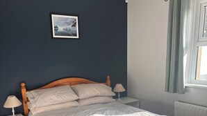Un dormitorio con una cama de madera, ropa de cama blanca, una mesita de noche con una lámpara, un cuadro enmarcado en la pared y una ventana con cortinas.