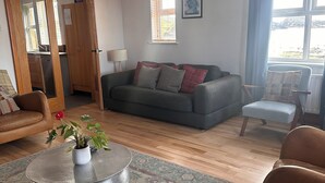 Una sala de estar con sofá, sillones, una mesa de centro con una planta, piso de madera y vistas al exterior a través de amplios ventanales.