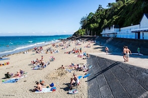 A crowded beach with people sunbathing and swimming, a lifeguard tower, and a building with a blue facade.