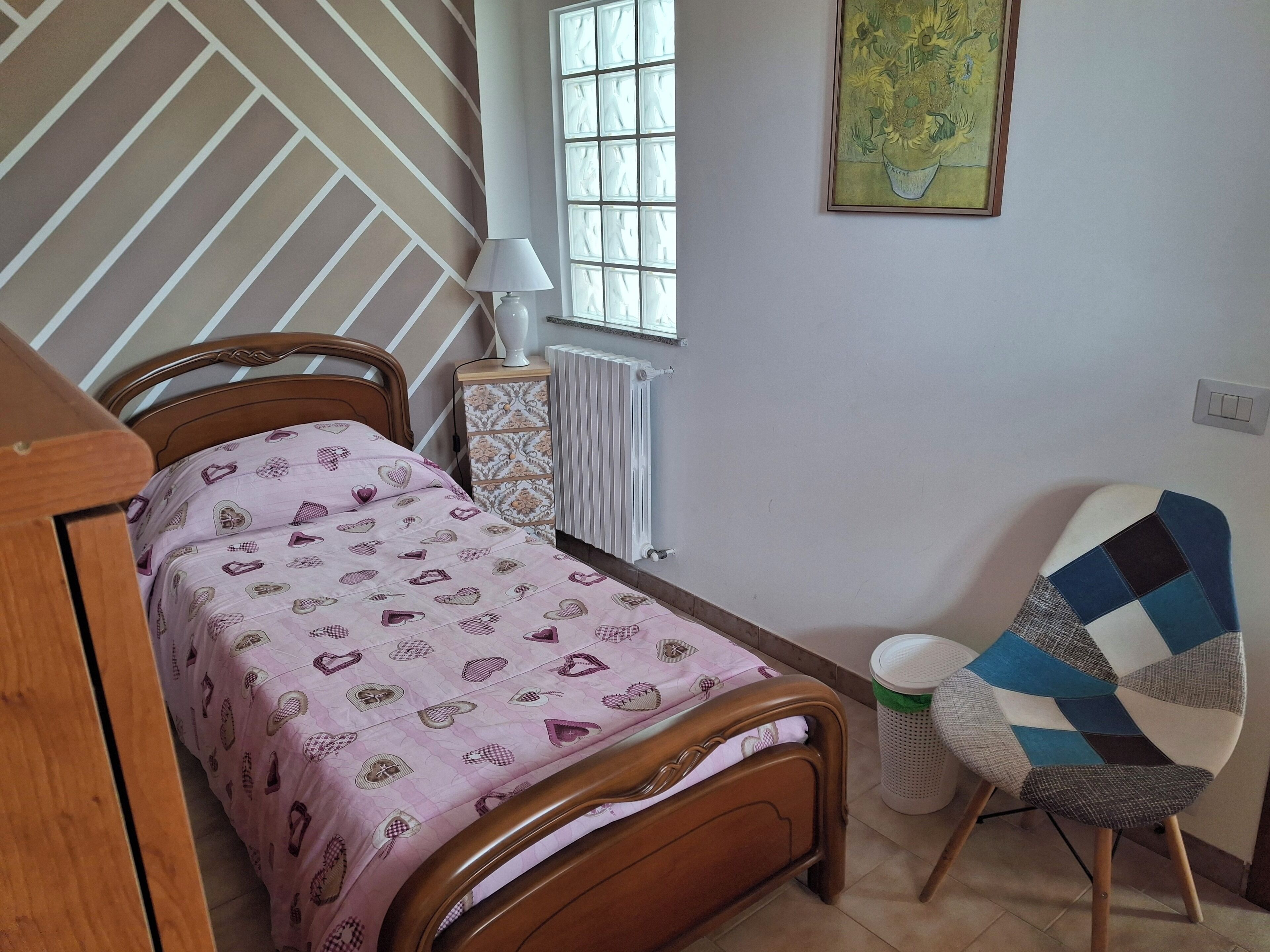 A bedroom with a bed, a radiator, a window, a lamp, and a chair.
