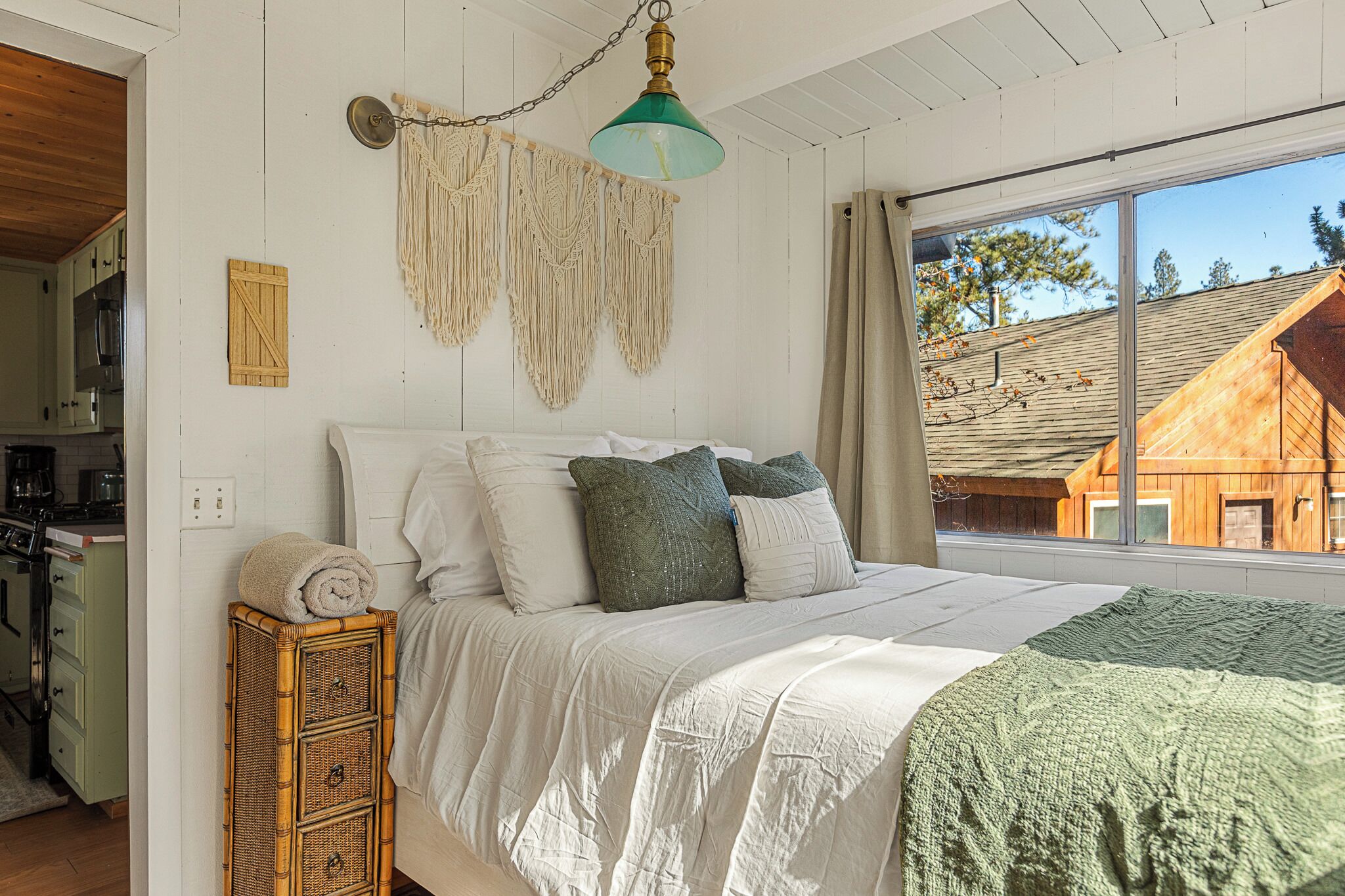 A bedroom with a bed, a hanging macrame light fixture, and a view of a wooden house through a window.