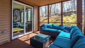A covered outdoor seating area with a large sectional sofa and a glass-top table, overlooking a forested landscape. - Rustic Ski Retreat! 2 min. walk to Ski Slopes, 6TVs, Linens, Wi-Fi, A/C, Firepit (bushkill)