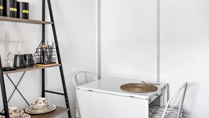 A compact room with a white table, a chair, a shelf with kitchenware, and a white door.