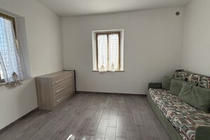 A room with a wooden dresser, a window with curtains, a patterned sofa, and a tiled floor.
