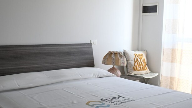 Uma cama bem arrumada com colcha branca, almofada com mensagem, abajur em uma mesa de cabeceira e janela com cortinas.