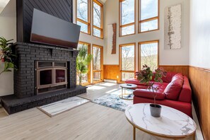 A spacious living room with a fireplace, large windows, a red sofa, and a marble coffee table. - Beautiful Modern Lodge! Pool Table,8 TVs Wi-Fi, Linen provided, Central AC (Saw Creek)