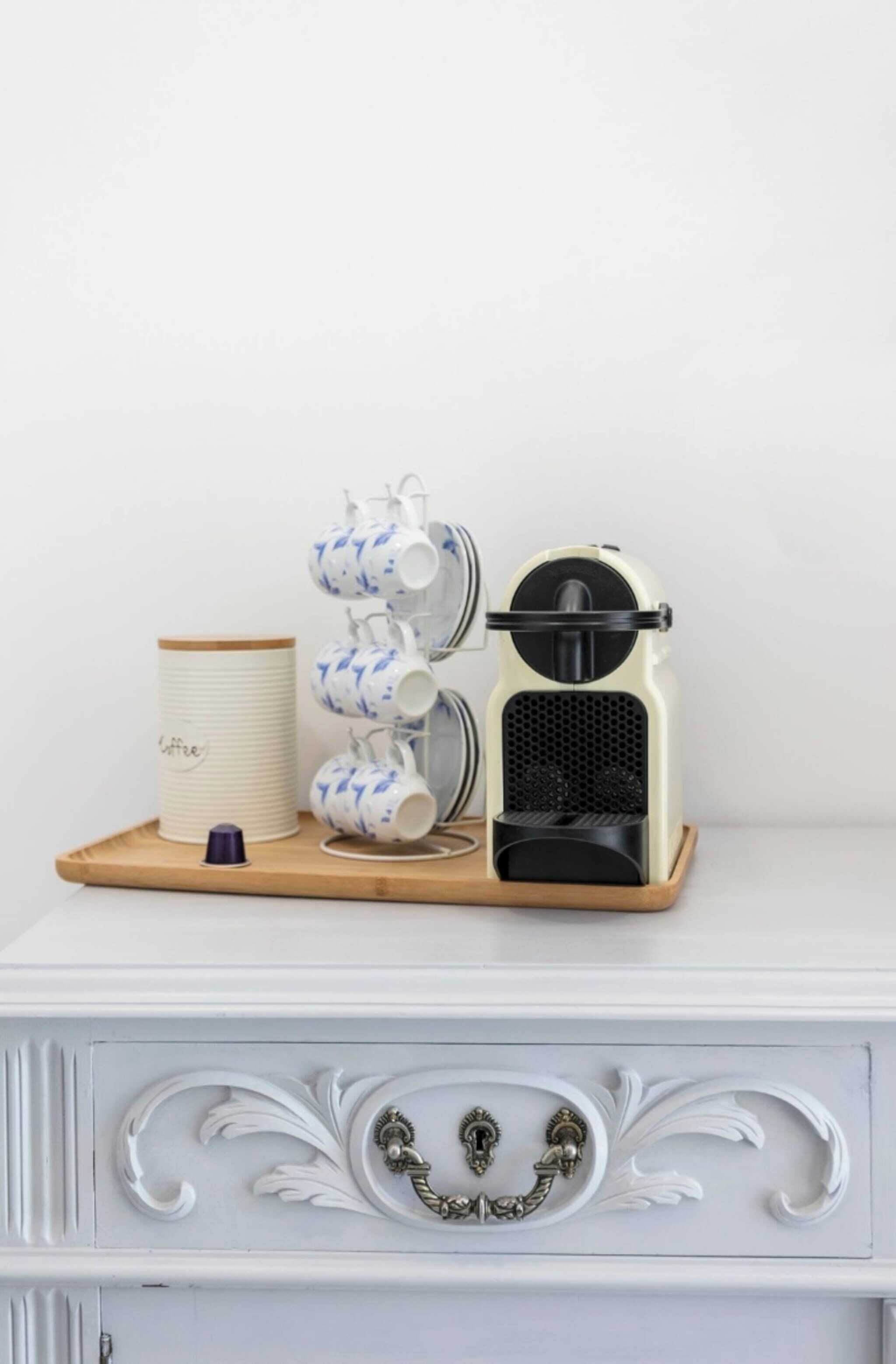 Uma máquina de café sobre uma bandeja, com uma pilha de chávenas e um recipiente sobre uma cómoda branca.