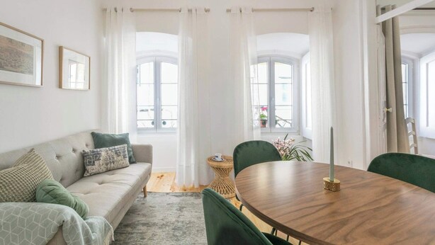 A living room with a sofa, a round wooden dining table, and two chairs. There are two windows with white curtains, a small side table, and a vase with flowers.