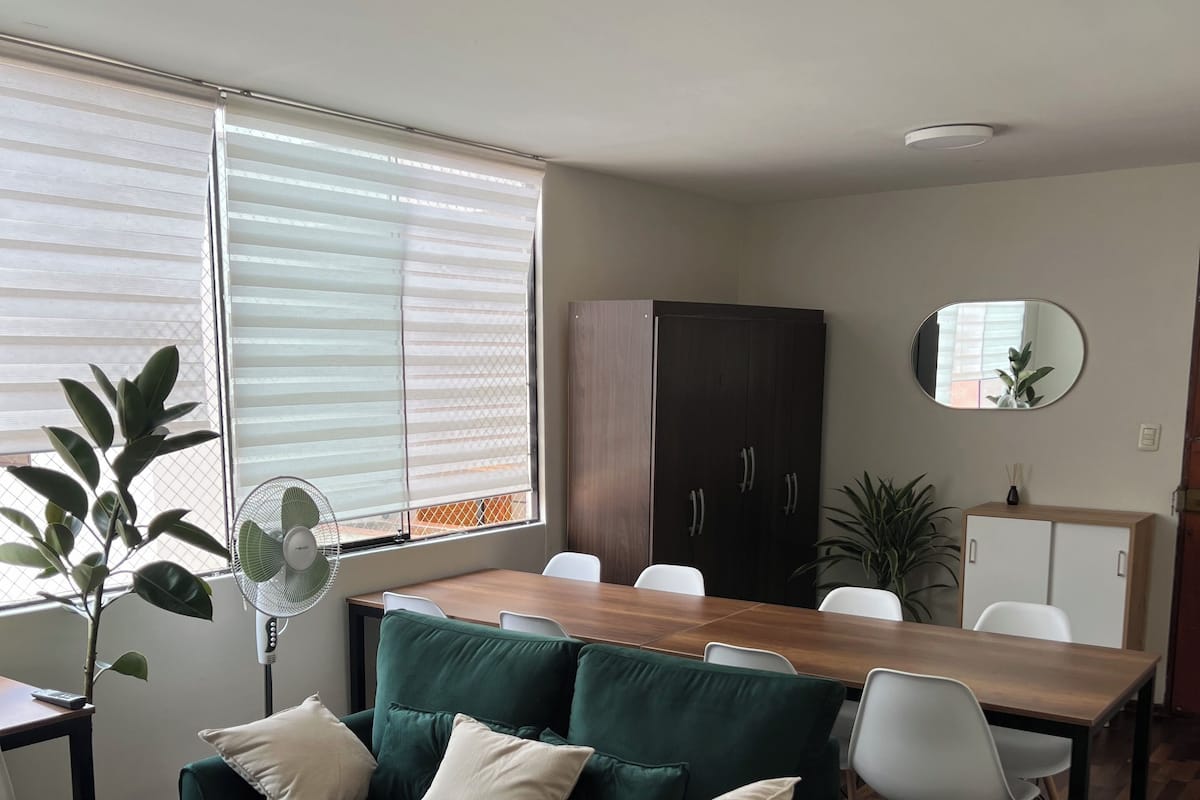 Un moderno salón con un sofá verde, una mesa de comedor de madera y sillas blancas. Hay una planta en maceta y un ventilador cerca de la ventana.