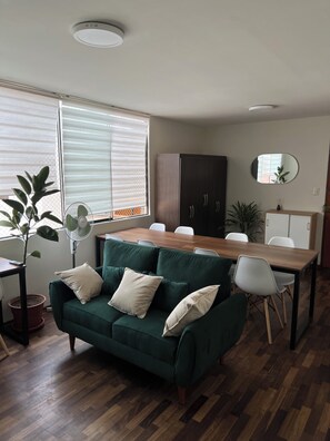 Un moderno salón con un sofá verde, una mesa de comedor de madera y sillas blancas. Hay una planta en maceta y un ventilador cerca de la ventana.