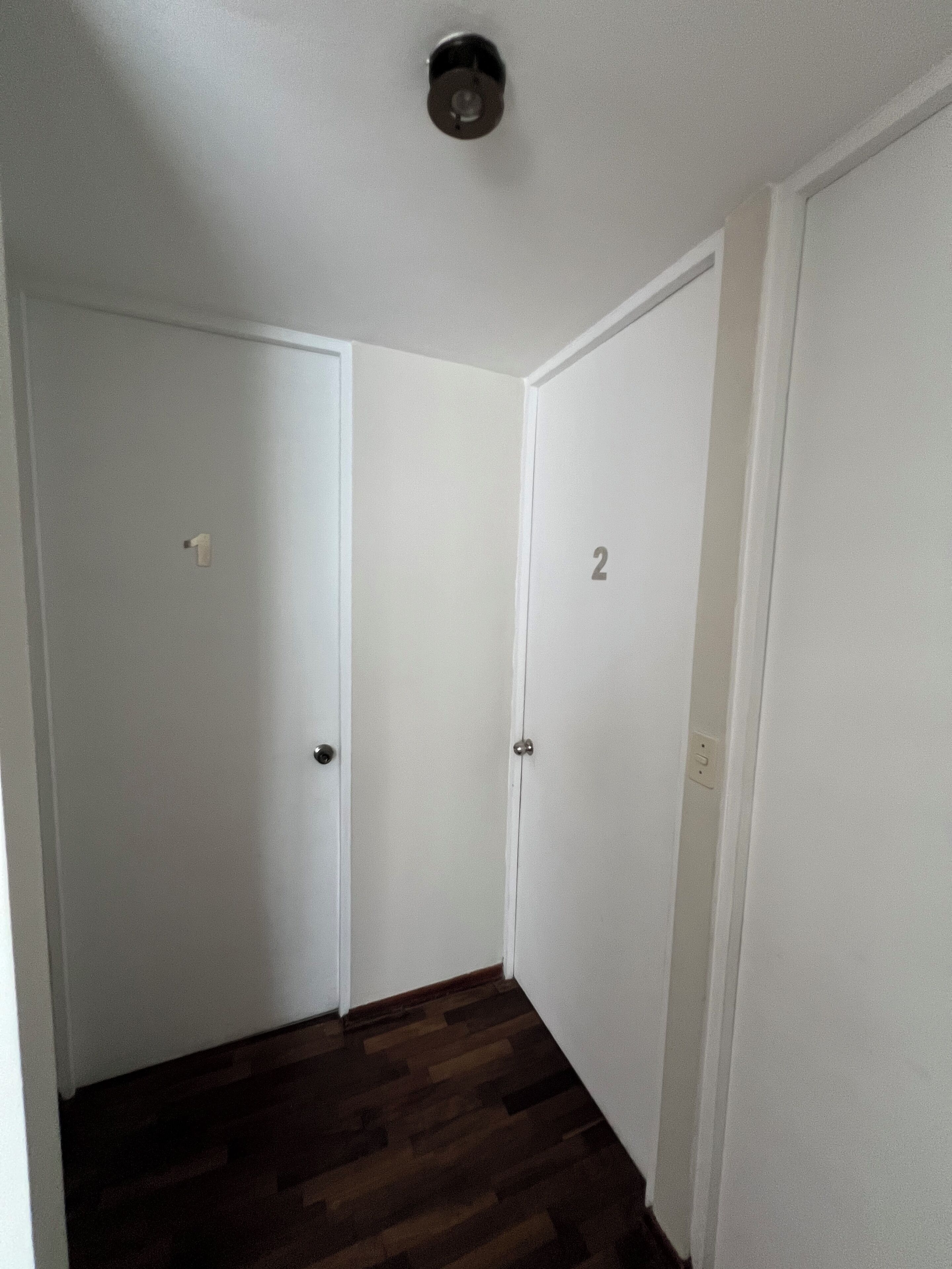 Un couloir étroit aux murs blancs et au plancher en bois. Il y a deux portes, l’une avec une poignée visible et l’autre portant le numéro « 2 ».