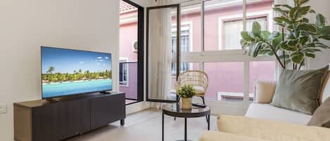 Una sala de estar con televisor, sofá y vista a un edificio rosa a través de un ventanal grande.