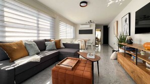A modern living room with a sectional sofa, a coffee table, and a wooden ottoman. There is a dining area in the background with a table and chairs.