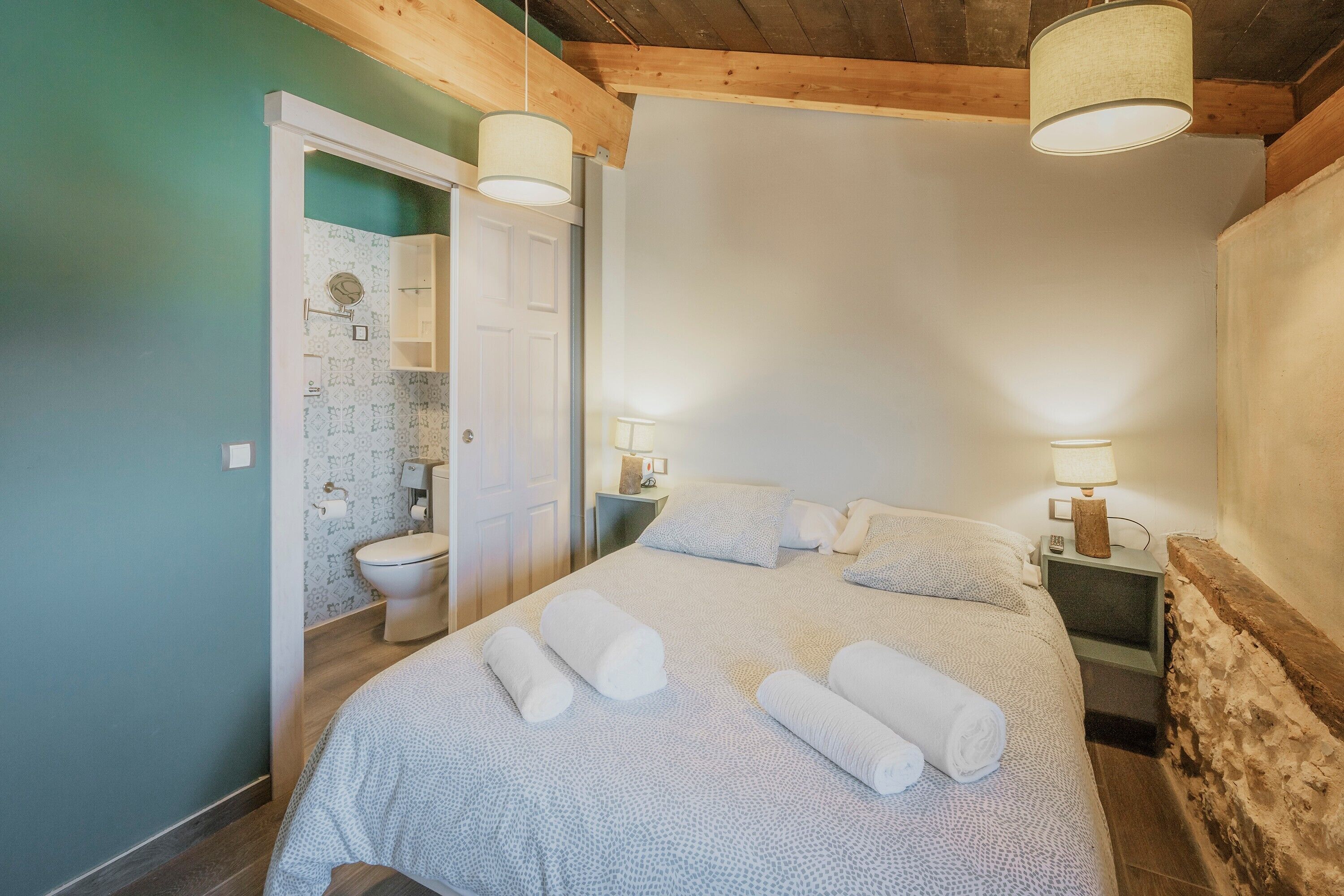 Quarto com uma cama, duas mesas de cabeceira, um criado-mudo, uma porta e um banheiro visível através de uma porta aberta.