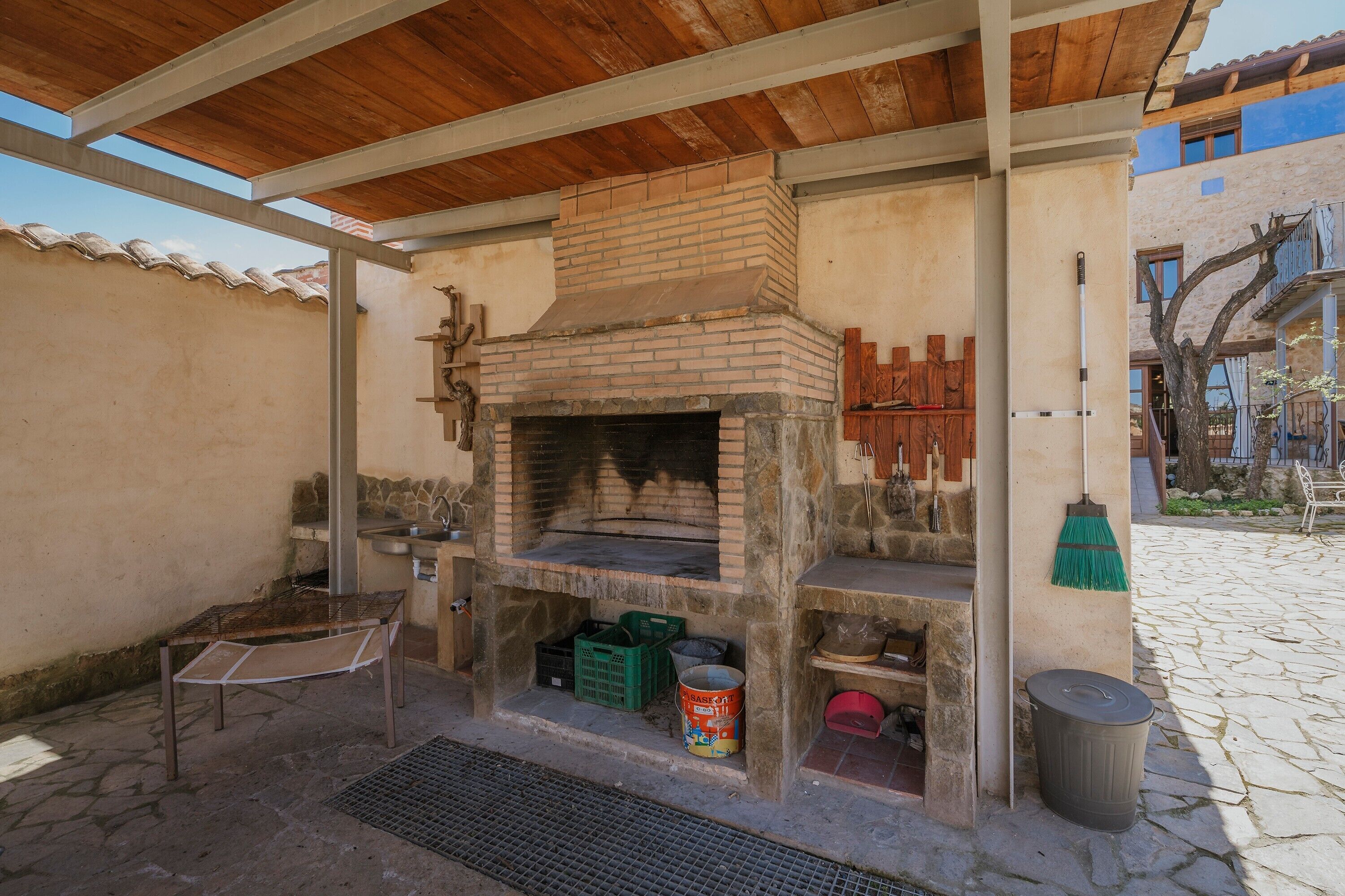 Un four en briques installé à l’extérieur, sous une pergola en bois, une table en métal et une poubelle.