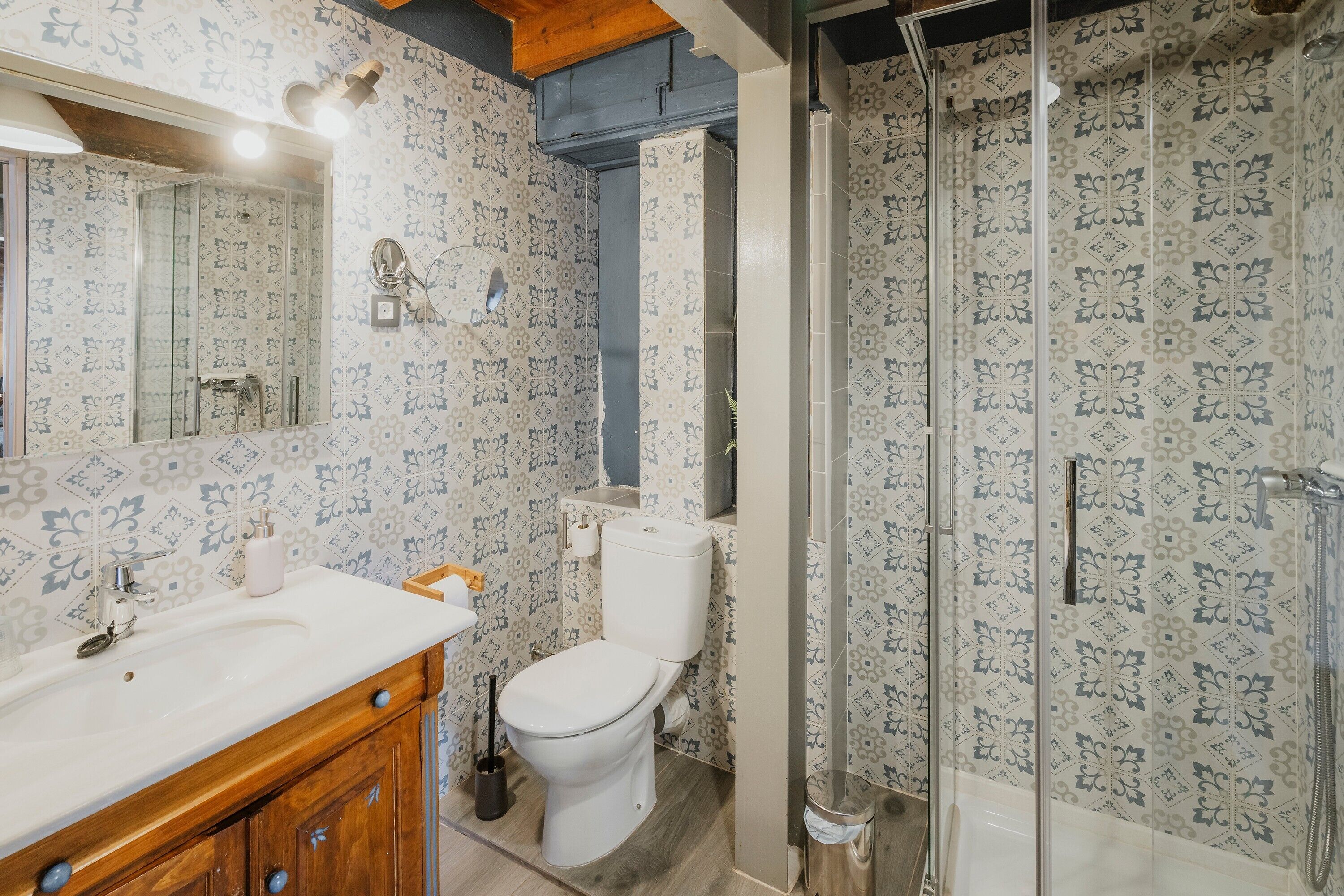 A bathroom with patterned wallpaper, a white sink, wooden cabinets, a toilet, and a glass-enclosed shower area.