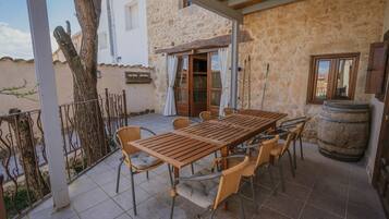 Un patio con una mesa de madera y sillas de mimbre, un edificio de piedra con una puerta, y un árbol.
