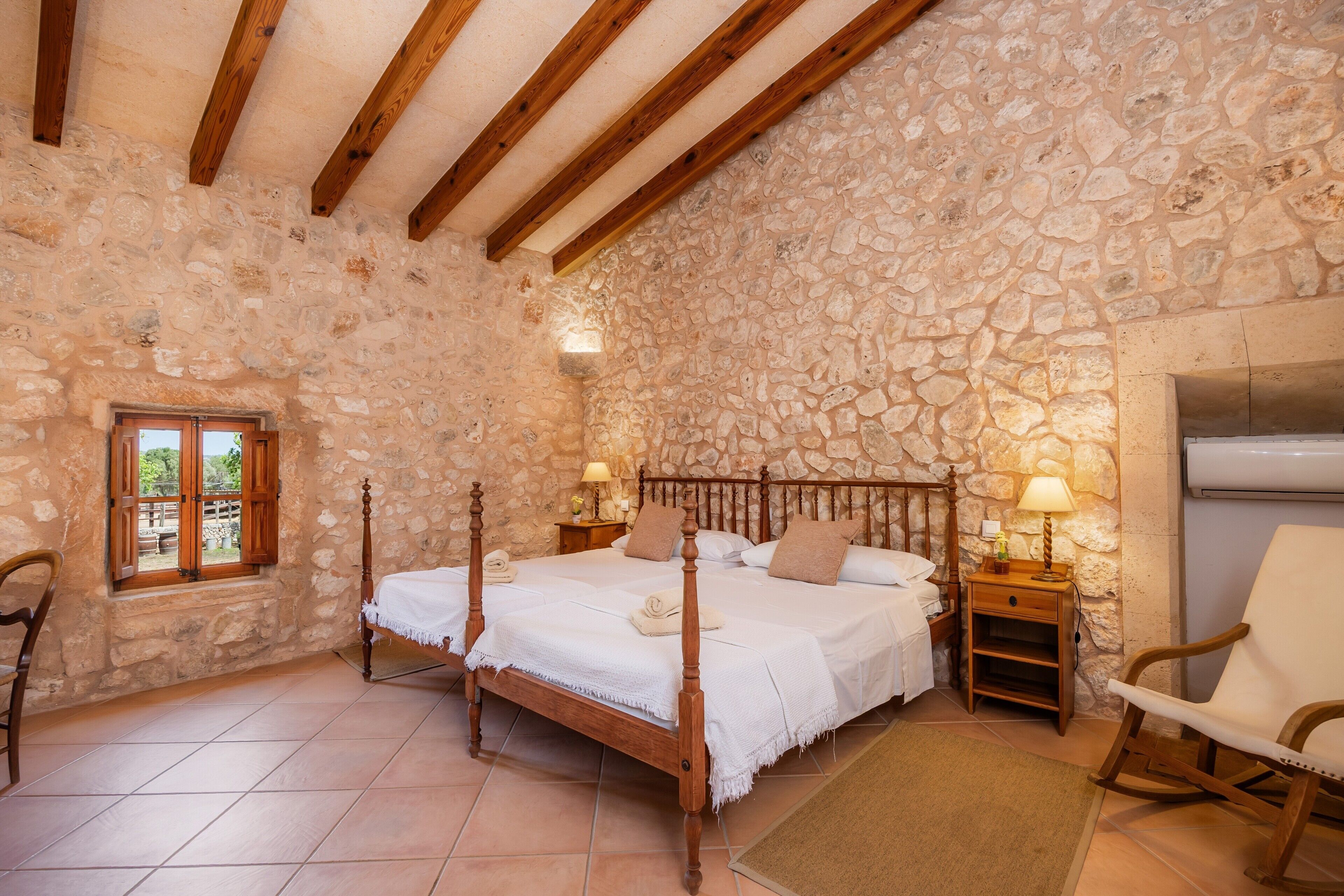 Une chambre aux murs de pierre, avec deux lits en bois, une fenêtre, une chaise et une table de chevet avec une lampe.