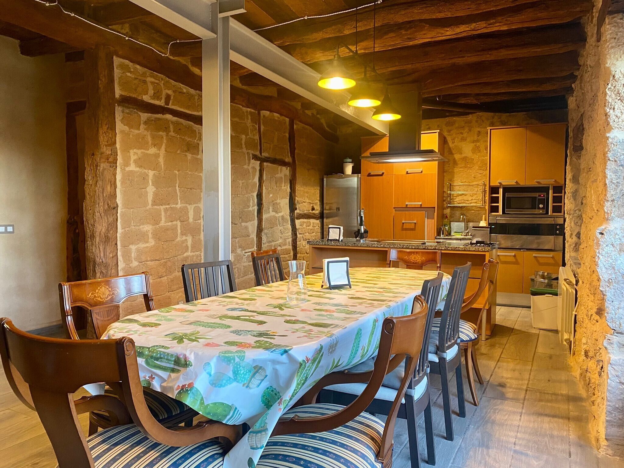 A rustic dining area with a long table, chairs, and a kitchen in the background.