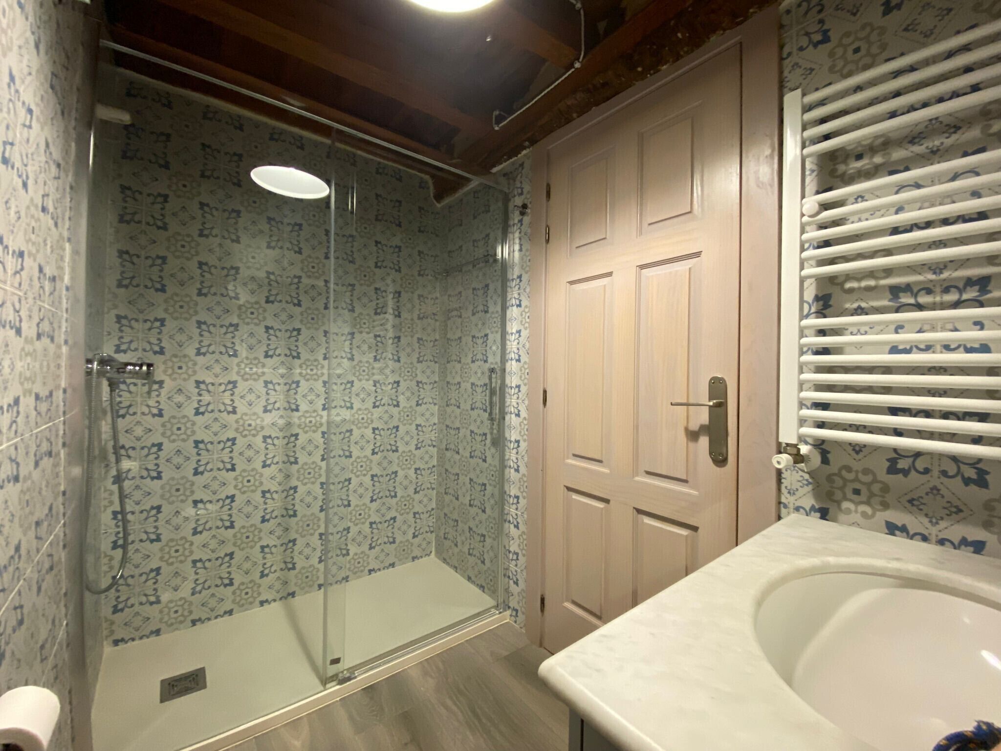 Un baño con baldosas estampadas, cabina de ducha con paredes de vidrio, puerta de madera y bañera blanca.