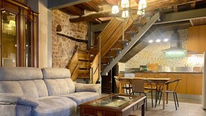 Een woonkamer met een bank, een salontafel met glazen blad, een eethoek met stoelen, een keuken met betegelde spatwand en een houten trap.