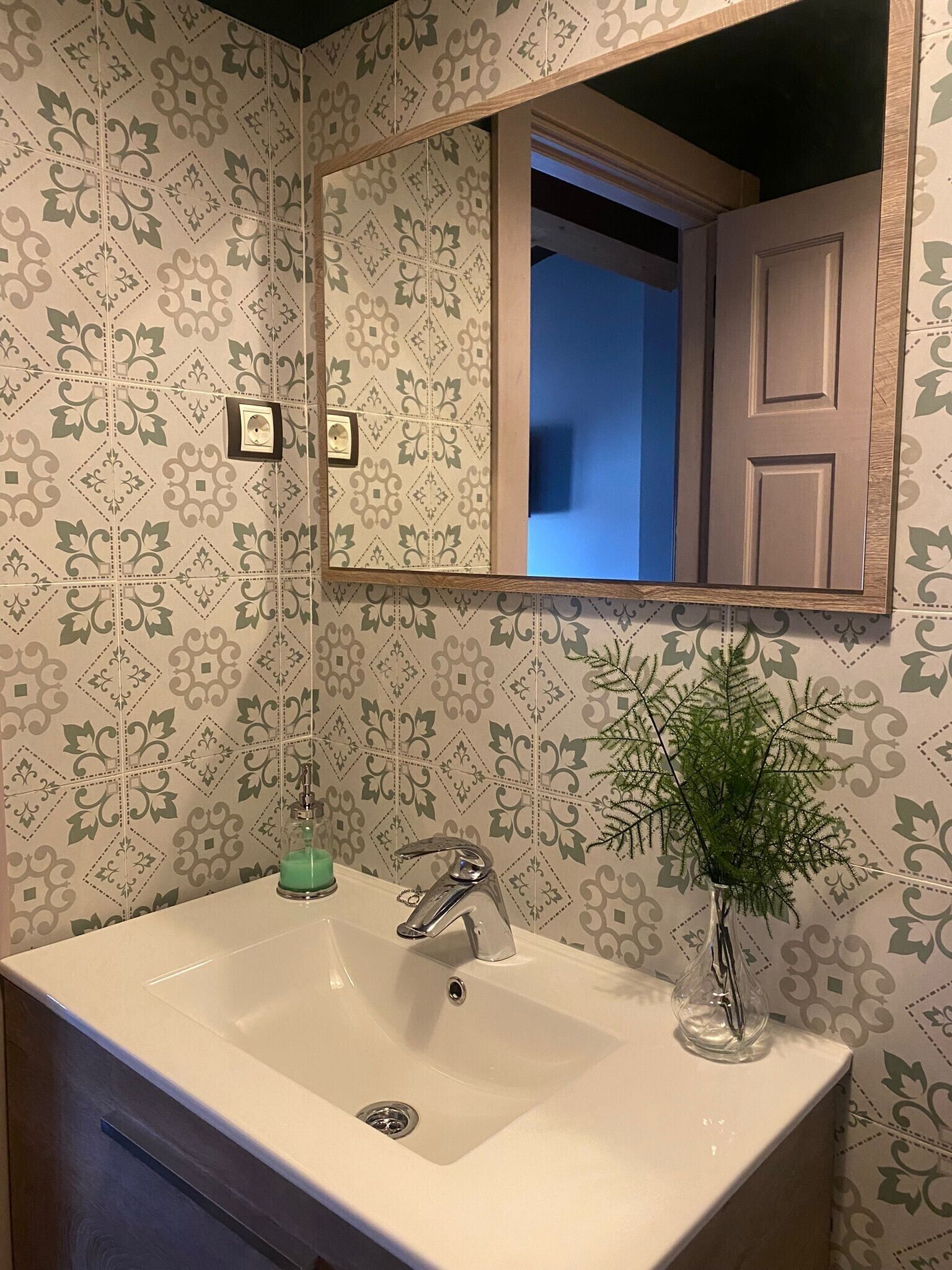 A bathroom with a white sink, a mirror, a green plant, and patterned wallpaper.