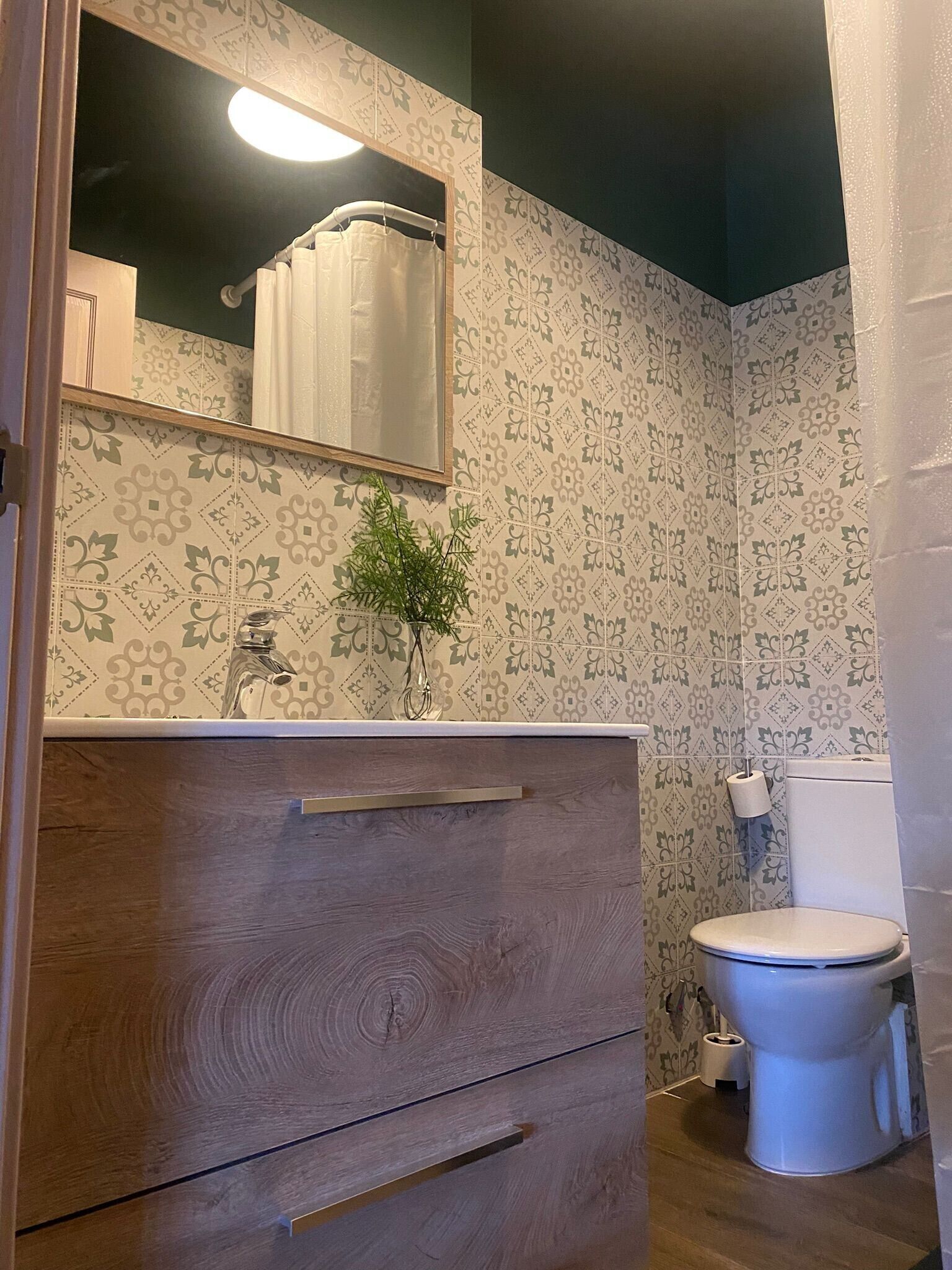A bathroom with patterned wallpaper, a wooden vanity, a mirror, a toilet, and a small plant.