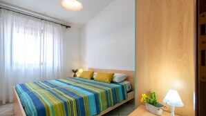 Una cama con un edredón a rayas de colores, una lámpara de noche y una planta pequeña sobre una mesita de noche de madera.