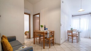 Una sala de estar con un sofá, una mesa de comedor y sillas. Hay una puerta que da a otra habitación.