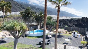 Zona de piscina con sillones reclinables, palmeras y montañas al fondo.