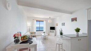 A modern kitchen with white cabinetry, a dining area with a table and chairs, a TV, and a view of the sea through a window.