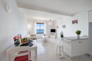 A modern kitchen with white cabinetry, a dining area with a table and chairs, a TV, and a view of the sea through a window.