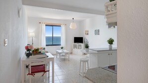 A modern kitchen with white cabinetry, a dining area with a table and chairs, a TV, and a view of the sea through a window.